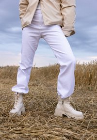 Botas beige con cremallera y suelas gruesas, combinadas con pantalones blancos de corte amplio, situadas en un campo con paja dorada. Se aprecian texturas suaves.