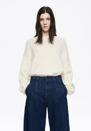 Suéter de punto en color crema claro con mangas anchas, combinado con pantalones vaqueros oscuros de cintura alta que cuentan con bolsillos frontales y costuras prominentes.