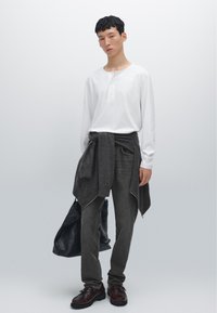 Weißes Langarm-Henley-Shirt, grauer Pullover um die Taille gebunden, graue Hose, braune Schuhe, hält eine schwarze Tasche. Einfaches, modernes Design.