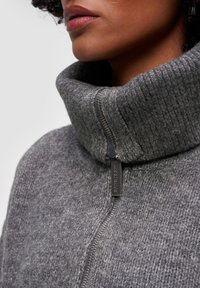 Szary wełniany sweter z wysokim prążkowanym kołnierzem, z zapięciem na zamek błyskawiczny i markowym uchwytem zamka. Miękka struktura, widok z bliska.