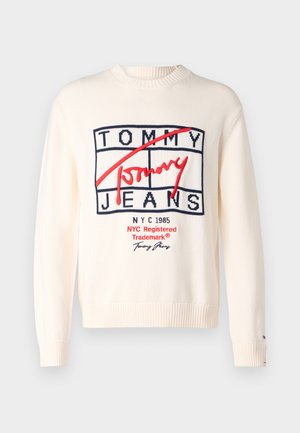 Krém színű kötött pulóver, amelynek középpontjában egy merész piros és tengerészkék logó áll, rajta a "TOMMY JEANS" és "NYC 1985" feliratokkal. Bordás mandzsetták és szegély.