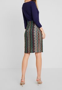 Kobieta ubrana w spódnicę w wielokolorowe paski do kolan, granatową bluzkę z długimi rękawami i beżowe sandały na wysokim obcasie, stojąca tyłem.