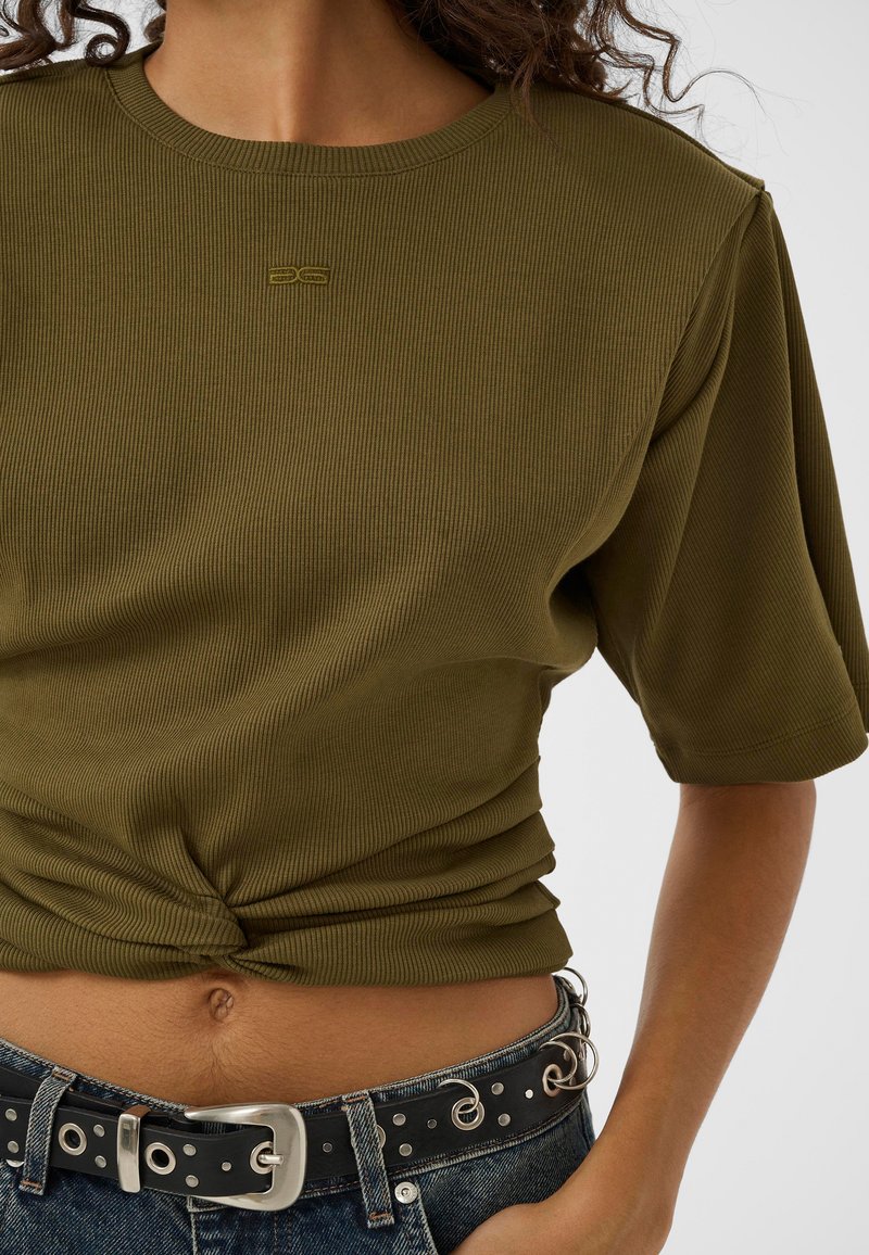 Femme portant un haut côtelé à manches courtes vert olive noué à la taille, avec un jean en denim foncé et une ceinture noire avec des œillets argentés.