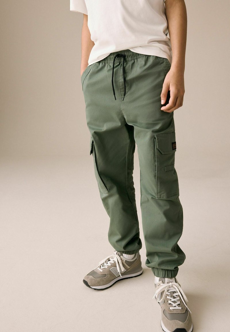 Pantalons cargo verts avec une taille élastique et des ourlets rabattus, dotés de poches latérales et d'une poche à patch sur la jambe. Portés avec des baskets grises.