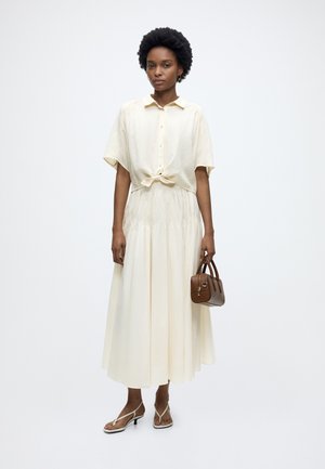 Femme aux cheveux courts naturels portant une chemise crème boutonnée nouée, une longue jupe crème, des sandales beiges et tenant un petit sac à main marron.