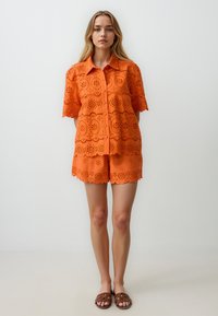 Oranje, oogjespatroon kortemouwen shirt en shorts set met geschulpte randen, gecombineerd met bruine instapsandalen.