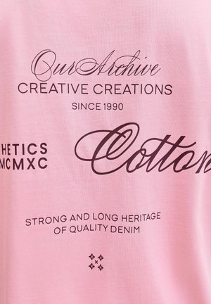 Roza bombažna majica s črnim tiskanim besedilom, ki vključuje "Naš Arhiv," "Kreativne Kreacije" in "Kakovostni Denim" v različnih pisavah.