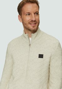 Hellbeige Zip-up-Pullover mit hohem Kragen, ausgestattet mit einem strukturierten diagonalen Muster und einem schwarzen Logopatch auf der linken Brust.