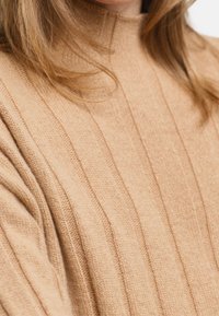 Maglione beige a collo alto a coste indossato, che mostra il collo e la spalla con ciocche di capelli castano chiaro sciolte.