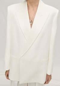 Blazer bianco oversize con rever affilati, due tasche frontali e una texture liscia. Una collana d'argento a più strati aggiunge un accento sottile.