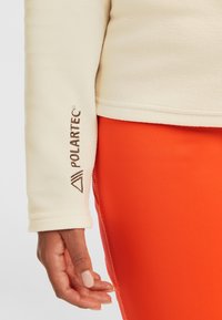 Kermä fleece-takki, jossa on Polartec-logo hihassa. Yhdistetty kirkkaan oranssiin kangastyyppiin, korostaen sileää tekstuuria ja kontrastia.