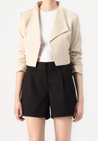 Beige croppet jakke med en bred krave og rynkede ærmer, kombineret med højtaljede sorte shorts. Glat tekstur og skræddersyet pasform.