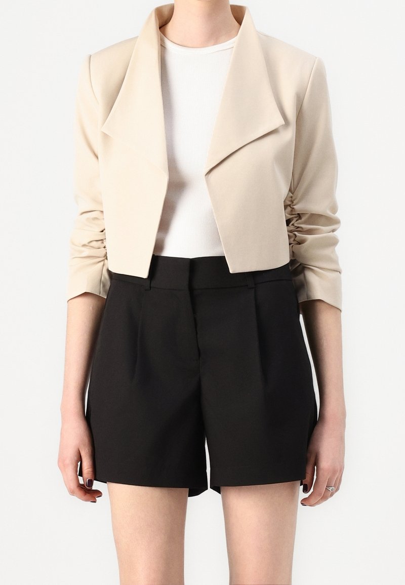 Beige croppet jakke med en bred krave og rynkede ærmer, kombineret med højtaljede sorte shorts. Glat tekstur og skræddersyet pasform.