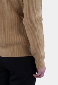 Beige ribbestrikket genser med lange ermer, teksturert stoff og en liten rød logo detalj nær kanten. Svart bukser synlig nedenfor.