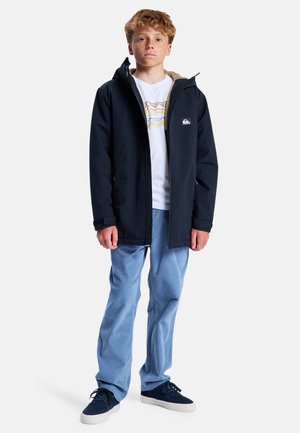 Garçon blond avec des taches de rousseur, portant une veste à capuche noire, un t-shirt blanc à motif graphique, un jean bleu clair et des baskets noires, debout devant un fond uni.