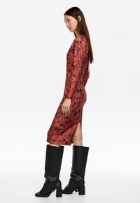 Vestido midi de manga larga en rojo oscuro con un patrón de salpicaduras negras. La tela parece suave y tiene una abertura lateral. Se combina con botas de cuero negras hasta la rodilla.
