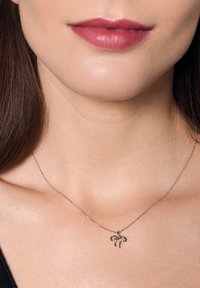 Srebrny naszyjnik z delikatnym wiszącym zawieszeniem w kształcie kokardy, umieszczony na gładkiej skórze dekoltu. Łańcuszek wydaje się cienki i subtelny.