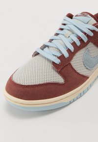 Sapatilha marrom e azul claro com parte superior em malha e têxtil, apresentando atacadores redondos, uma sola azul clara e um swoosh na lateral.