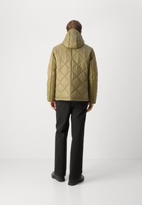 Veste matelassée en vert olive avec capuche, présentant des motifs en diamants et des poignets élastiques, portée sur un pantalon noir et des chaussures.