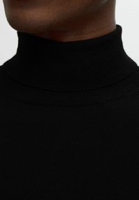 Svart polotröja med ribbad textur, kroppsnära design och hög krage. Materialet verkar mjukt och elastiskt.