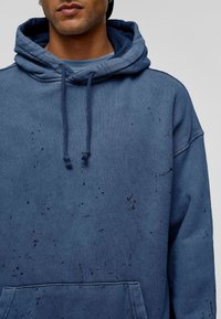 Blauwe hoodie van zacht materiaal met een voorzak en trekkoorden. Heeft subtiele zwarte spikkels verspreid over de hele hoodie.