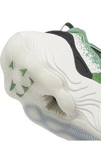 Gros plan sur la semelle d'une chaussure de sport blanche et verte, présentant une surface antidérapante texturée, des crampons triangulaires verts et un clip talon noir.