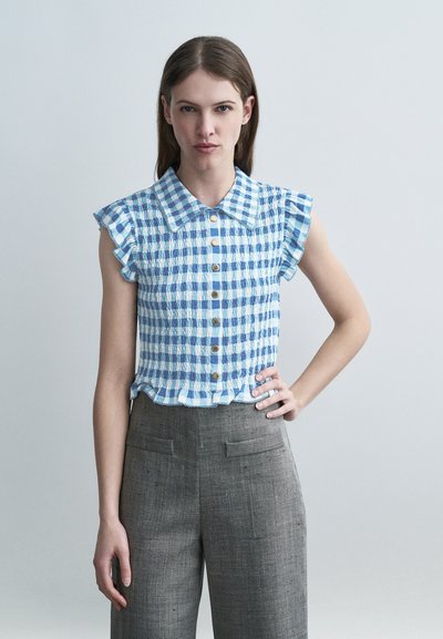 Femme portant un chemisier sans manches à carreaux bleus et blancs avec des boutons et un pantalon gris taille haute, debout avec une main sur la hanche.