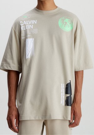 Beżowy, oversize'owy t-shirt Calvin Klein Jeans z abstrakcyjnymi grafikami w biało-czarnym i zielonym motywie kosmicznym, noszony przez osobę.