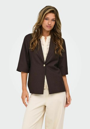 Vrouw met lang golvend haar die een donkerbruine blazer draagt over een witte kanten blouse en lichtbeige broek, staand tegen een effen lichte achtergrond.
