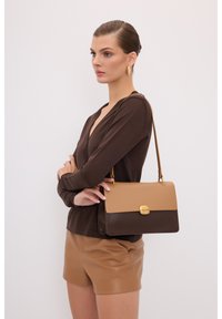 Borsa a mano in pelle marrone con una patta color crema, hardware dorato e tracolla. Il corpo è di una tonalità più scura, con una texture liscia.