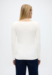 Γυναί κα με ίσια ξανθά μαλλιά, φορώντας εφαρμοστή λευκή μπλούζα με μακριά μανίκια και σκούρο μπλε τζιν, εμφανιζόμενη από την πλάτη μπροστά σε απλό φόντο.