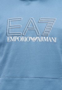 Niebieska bluza z kapturem z dużym białym logo "EA7 EMPORIO ARMANI" i paskowanymi akcentami. Materiał wydaje się miękki i teksturowany.