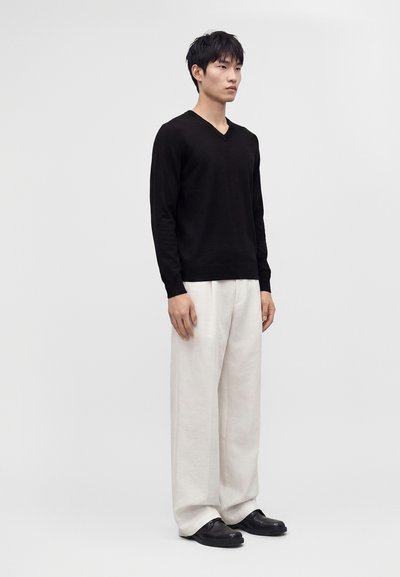 Jeune homme debout face à l’objectif, portant une chemise noire à manches longues, un pantalon blanc ample et des chaussures noires sur un fond blanc uni.
