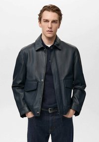 Dunkelblaue Lederjacke mit Reißverschluss, Punktkragen, zwei vorderen Taschen und glatter Textur, kombiniert mit einem schwarzen Hemd und dunklen Jeans.
