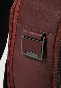 Braune Ledertasche mit strukturierter Oberfläche, Metallbeschlägen mit Logo und einem Reißverschlussdetail an der Seite.