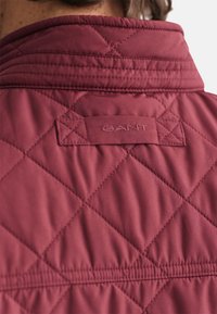 Bordeaux gewatteerde jas met een staande kraag, voorzien van een gestikt patroon en een textuurlabel met "GANT" aan de achterkant.