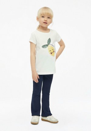 Enfant blond portant un t-shirt blanc avec un citron en sequins et des feuilles vertes, pantalon bleu marine et baskets blanches, debout avec une main sur la hanche.