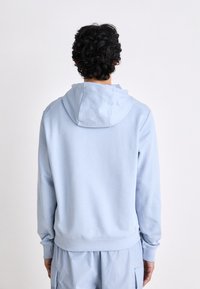 Personne aux cheveux bouclés foncés portant un sweat à capuche bleu clair et un pantalon assorti, debout face à un fond blanc uni.