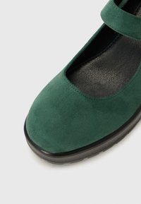 Zaļas zamša Mary Jane kurpes ar noapaļotu purngalu un siksniņu pāri augšdaļai, novietotas uz gaiša fona.