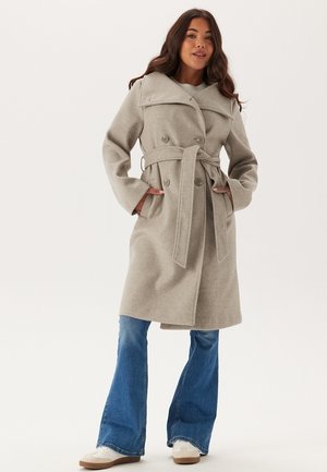 Trench coat grigio chiaro in misto lana con ampio colletto, bottoni doppi e vita con cintura. Indossato sopra jeans a zampa.