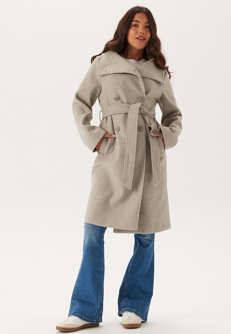 Trench coat grigio chiaro in misto lana con ampio colletto, bottoni doppi e vita con cintura. Indossato sopra jeans a zampa.