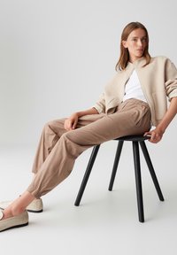 Beige zip-up jacket over a white top, paired with light brown elastic waist pants. Smooth fabric with a relaxed fit, featuring a black stool.