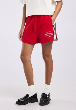 Persoon met rode shorts met elastische tailleband, witte zijstrepen en tekst, witte sokken, zwarte loafers en een loszittend wit bovenstuk, handen in de zakken.
