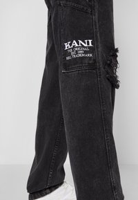 Pantalones de mezclilla negra con detalles desgastados, que presentan un logo y texto bordados en blanco en el bolsillo lateral izquierdo.