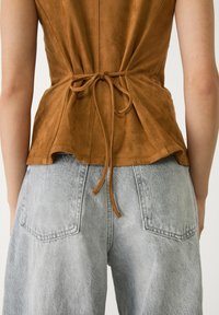 Bruine suède top met een getailleerde taille en een strikdetail aan de achterkant, gecombineerd met vervaagde grijze wijde denimbroek.