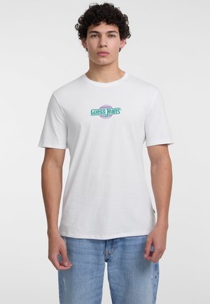 Bijela pamučna majica s kratkim rukavima koja na sredini ima zeleni i ljubičasti logo "Guess Jeans". Labav kroj i okrugli izrez.