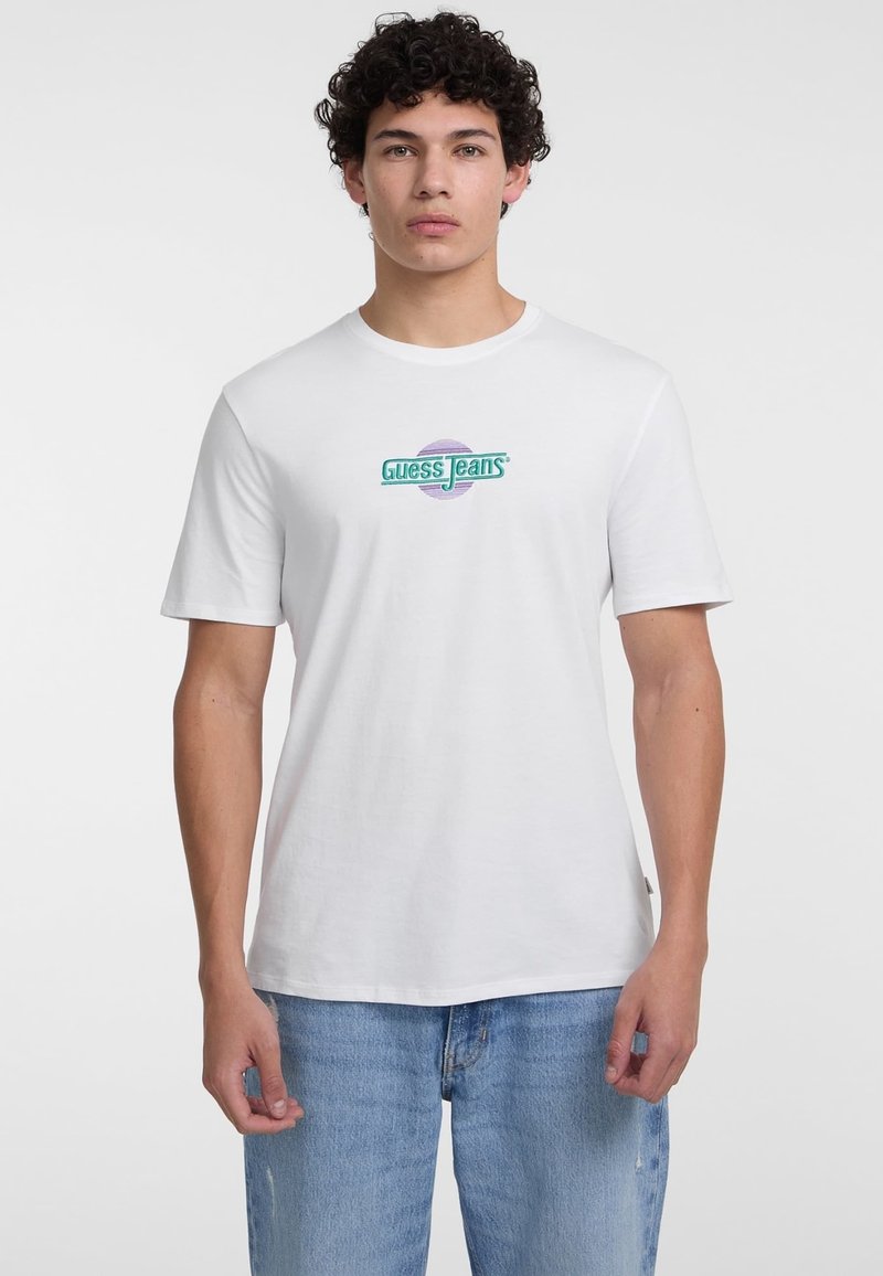 Baltas medvilninis marškinėliai su žaliu ir purpuriniu "Guess Jeans" logotipu centre. Trumpomis rankovėmis, laisvu kirpimu ir apvalia iškirpte.