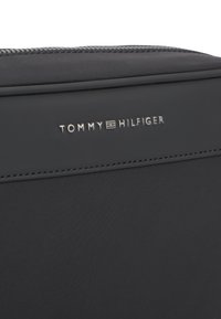Κοντινό πλάνο μαύρης υφασμάτινης τσάντας με ασημί λογότυπο «Tommy Hilfiger» και φερμουάρ κατά μήκος της κορυφαίας ακμής.