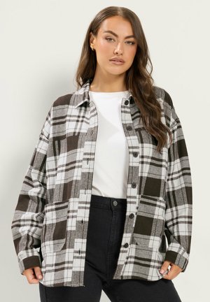 Femme portant une chemise à carreaux noire et blanche boutonnée sur un haut blanc et un pantalon noir, se tenant devant un fond clair uni.
