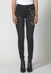 Kvinna som bär svarta skinny jeans med blommig broderi på höger lår och svarta ankelstövlar, stående mot en enfärgad bakgrund.